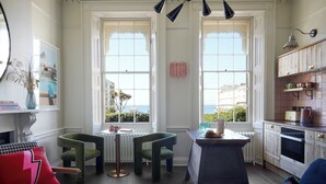 A cozy room with a view of the sea, featuring a dining table, two chairs, a sofa, and a kitchen area with cabinets and a radiator. - Artist Residence Brighton (Brighton)