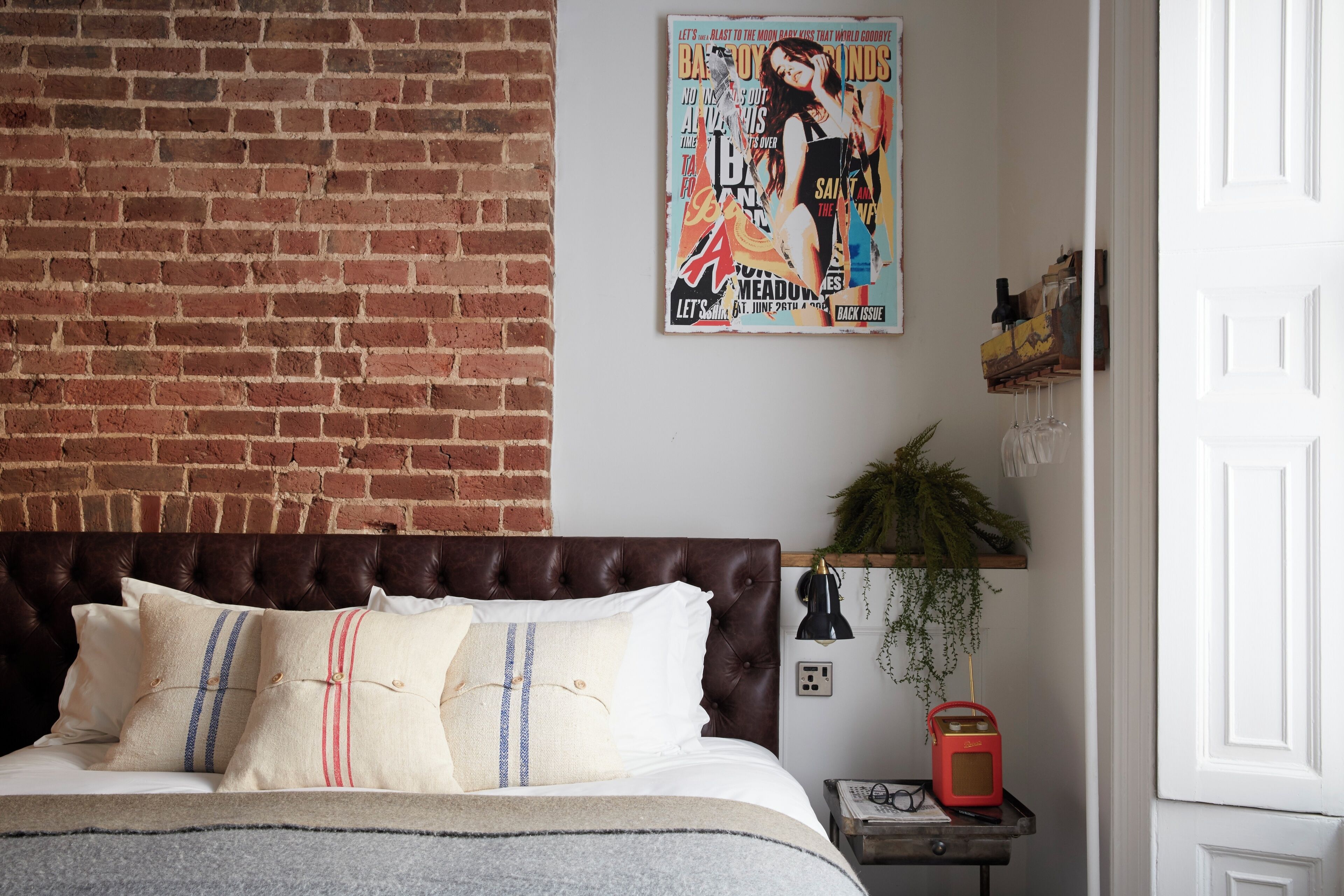 A bedroom with a brick wall, a bed with striped pillows, a magazine on the wall, a small table with a lamp, and a door.