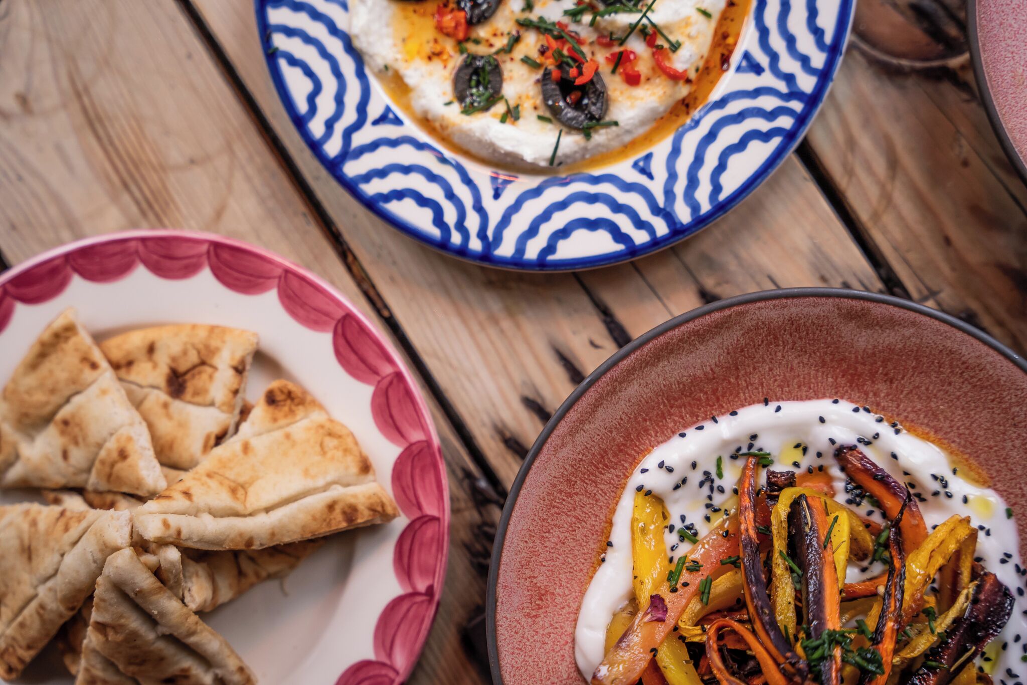 Une table en bois sur laquelle se trouvent trois assiettes de nourriture : une avec du pain plat, une avec une sauce blanche et des légumes, et une autre avec une sauce rouge et des légumes.