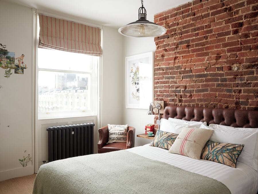 Un dormitorio con pared de ladrillo, una cama grande, una ventana con persianas, un radiador y una silla con cojín.