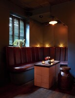 Una habitación tenuemente iluminada con un banco de madera, una mesita con velas y una planta en maceta junto a una ventana con persianas.