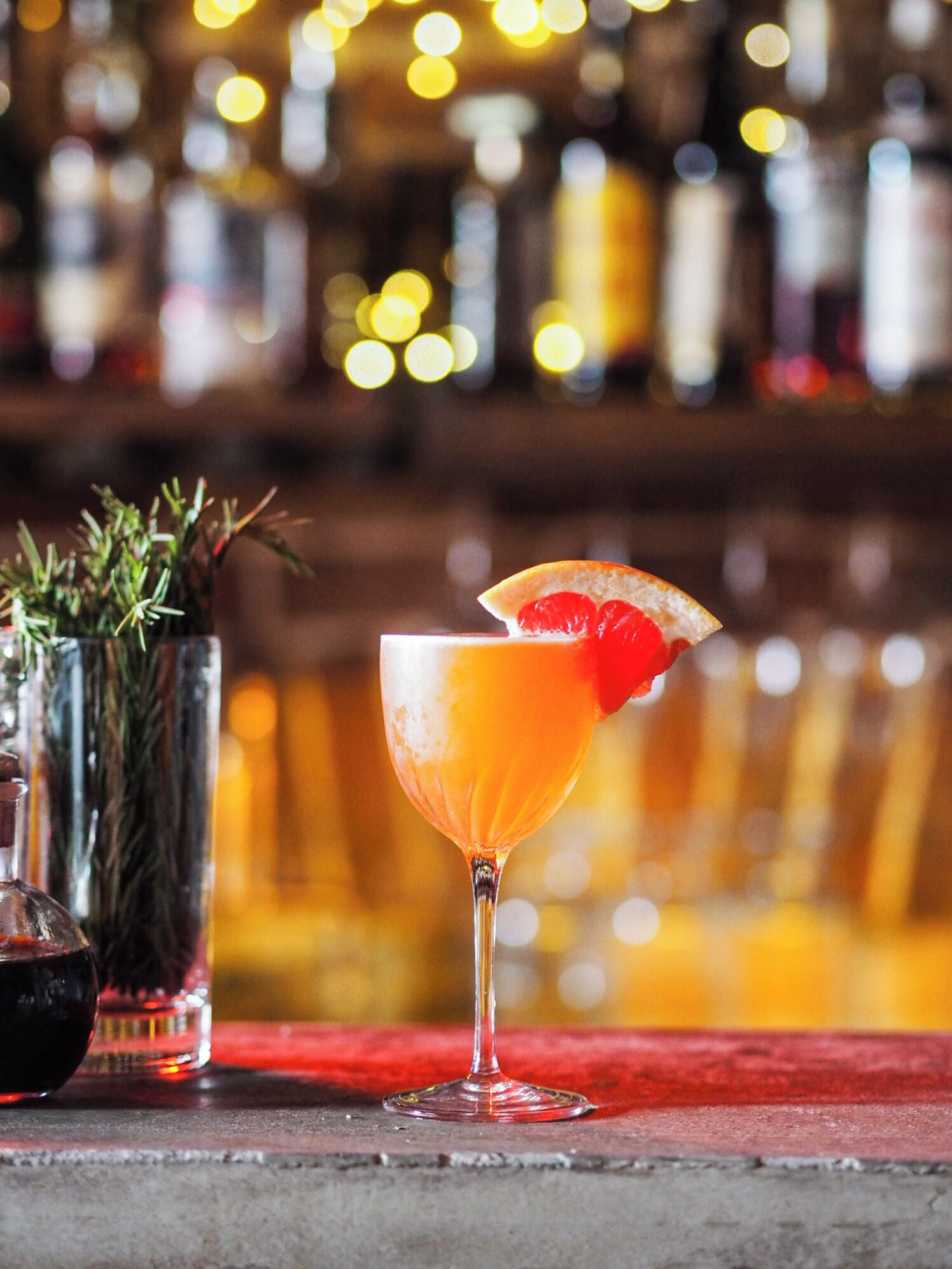 A cocktail in a stemmed glass with a slice of orange and a cherry on the rim, placed on a bar counter with a blurred background of bottles and lights.