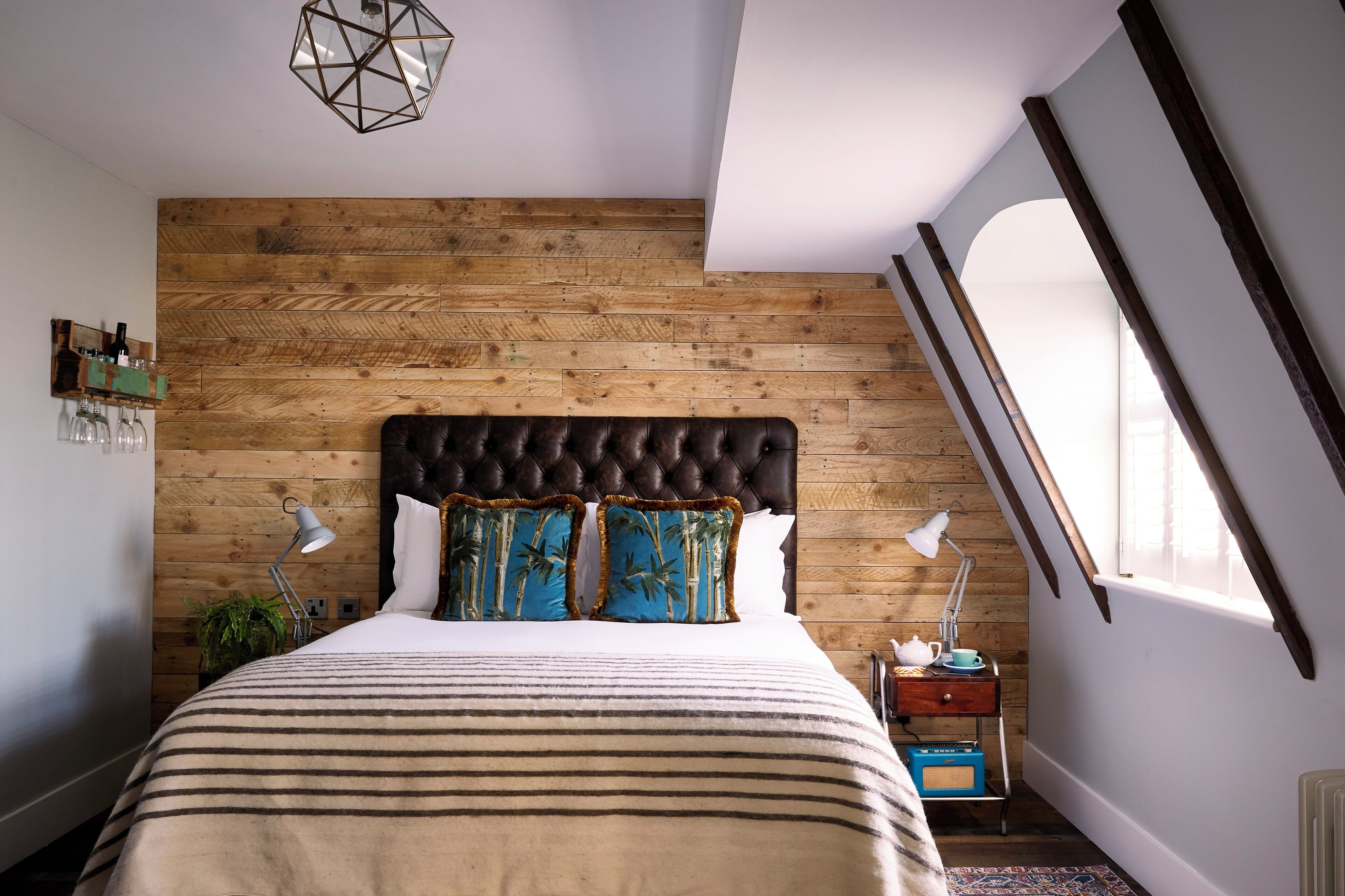 Ein Schlafzimmer mit einem Holzkopfteil, einem Bett mit gestreifter Bettwäsche, zwei Zierkissen, einem Nachttisch mit Lampe und einem Fenster mit Blick.