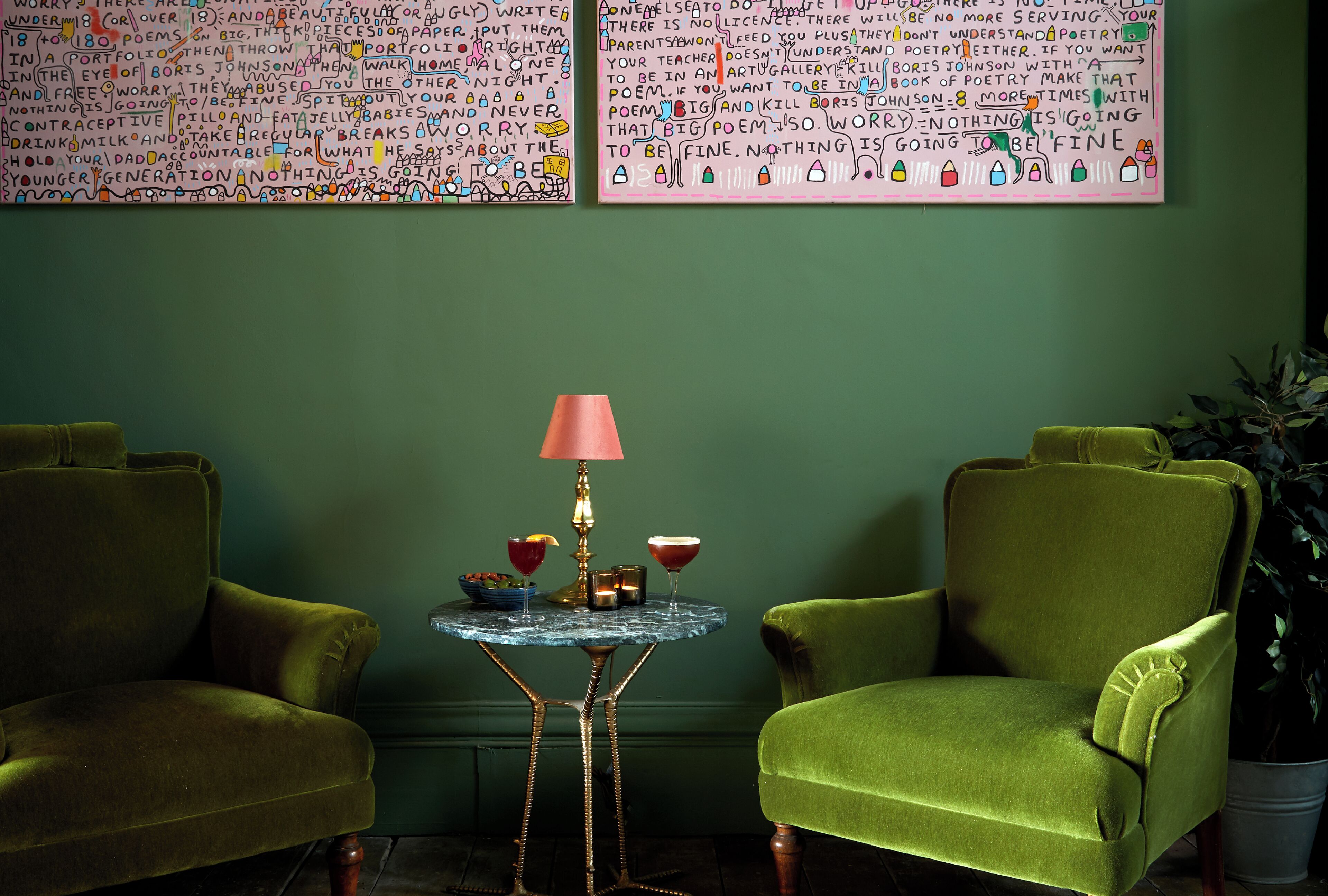 A room with two green armchairs, a small table with a lamp, and two large pieces of wall art.