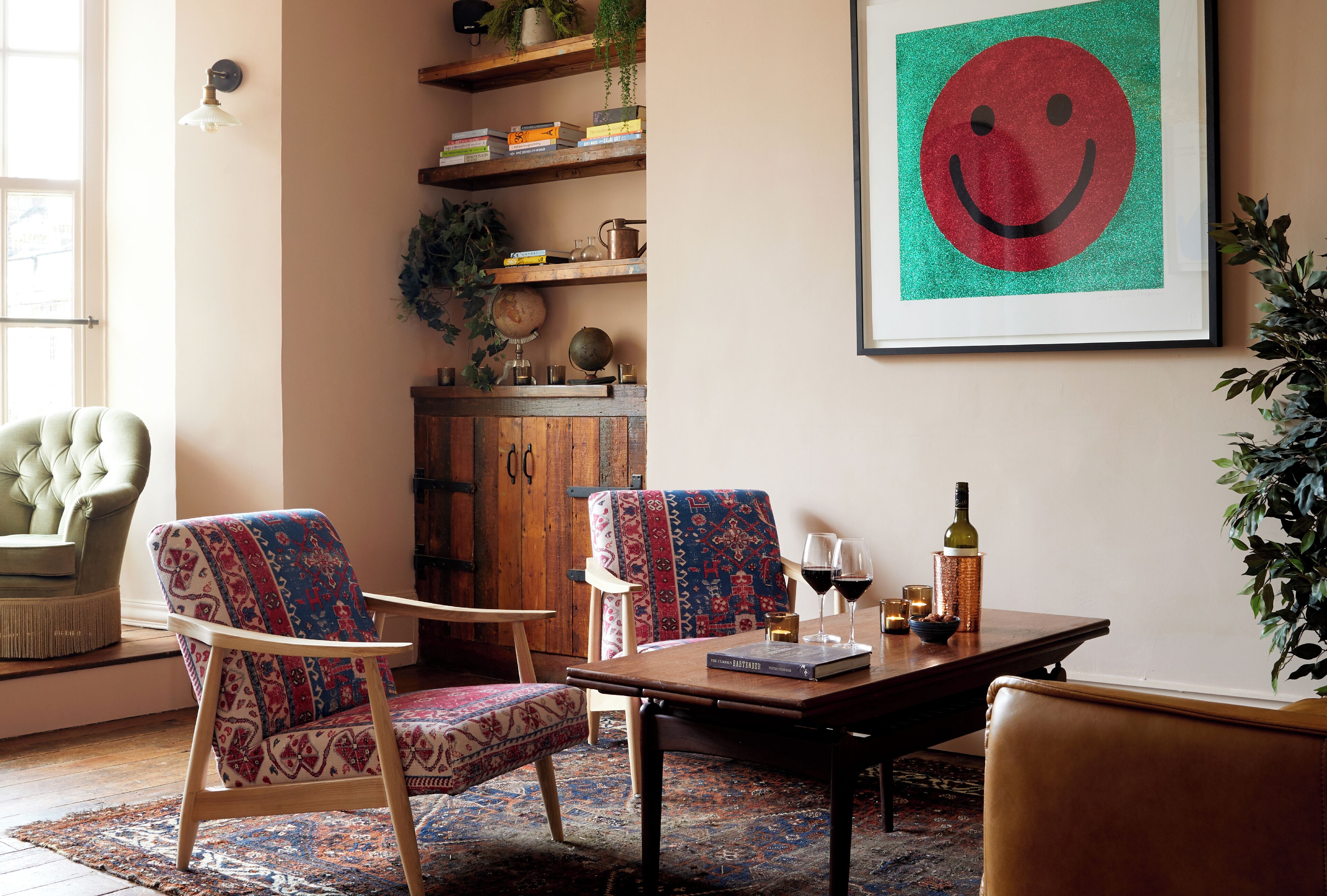 A cozy living room with a wooden coffee table, two patterned armchairs, a bookshelf, and a framed smiley face artwork.