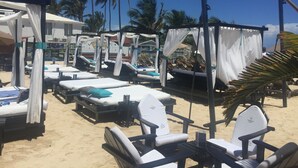 Un hôtel situé en bord de mer, doté de nombreuses chaises longues et de parasols, entouré de palmiers et surplombant un ciel bleu azur.