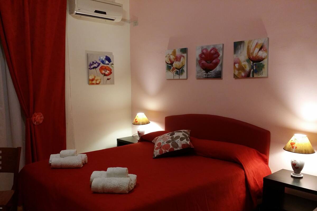 A hotel room with a red bed, two bedside lamps, a red curtain, and three floral paintings on the wall.