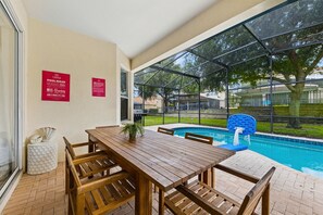 A poolside area with a wooden table and chairs, a pool, and a glass enclosure.
