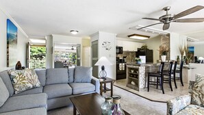 A modern living room with a grey sofa, a wooden coffee table, and a kitchen area with a dining set. - Oceanfront 2bd, lanai with Kaanapali pool views (Lahaina)