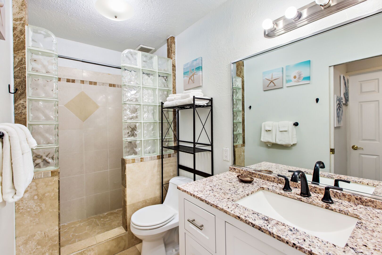 A bathroom with a glass shower enclosure, a toilet, a sink with a granite countertop, and a mirror with artwork.