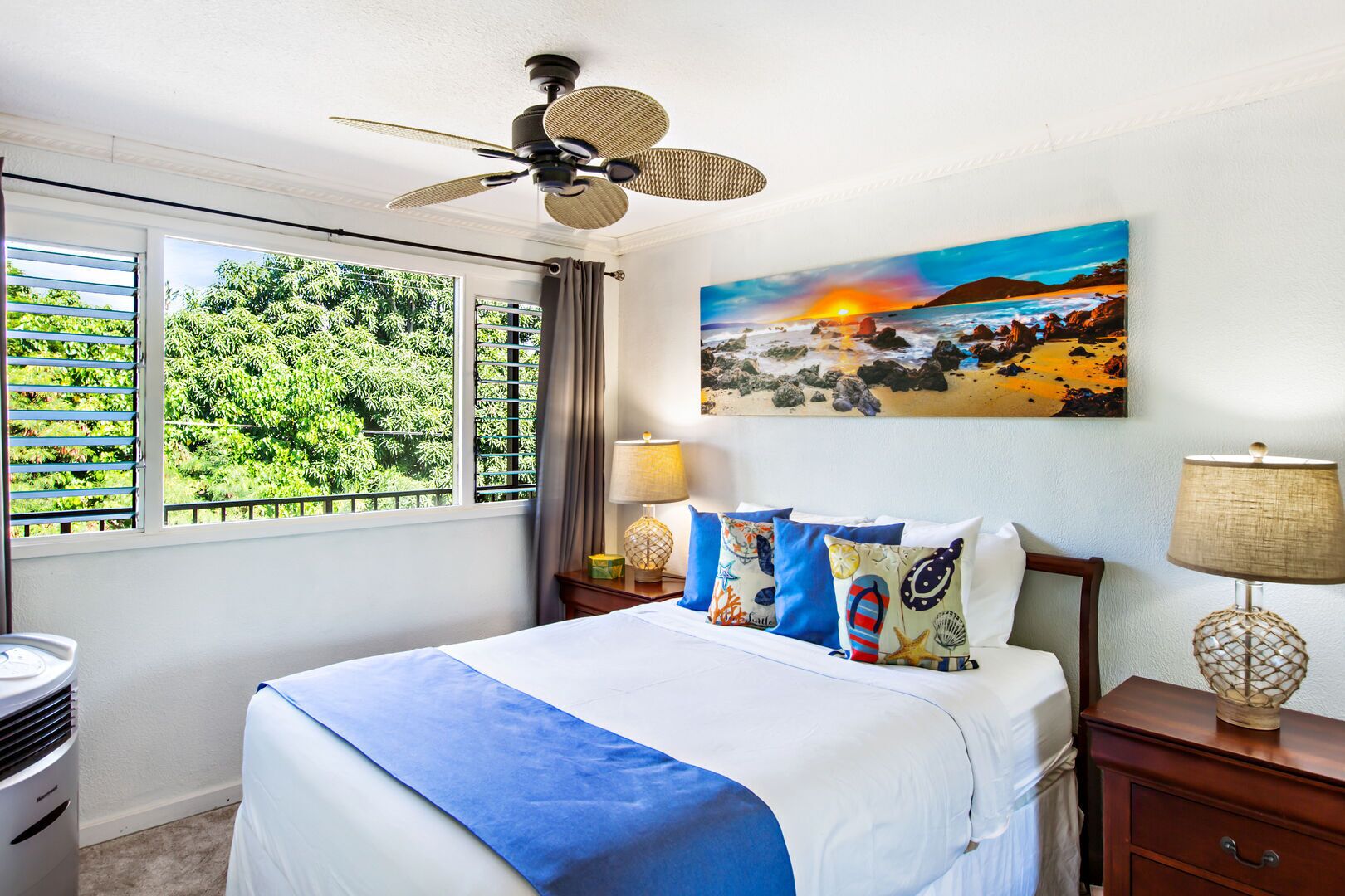 Een hotelkamer met een bed, een plafondventilator, een schilderij van een zonsondergang en uitzicht op groen buiten het raam.