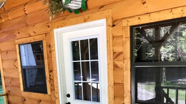 Una cabaña de madera con puerta blanca, dos ventanas, una pequeña mesa con sillas y una planta en maceta.