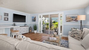 Ein Wohnzimmer mit einer Couch, einem Couchtisch, einem Fernseher, einer Küchenzeile mit Arbeitsfläche und Schränken sowie einer Glasschiebetür, die auf eine Terrasse führt.