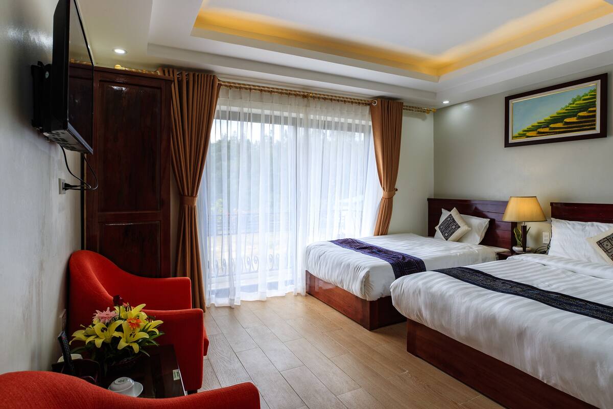 Ein Hotelzimmer mit zwei Betten, einem an der Wand befestigten Fernseher, einem Schreibtisch mit Stuhl und einem Fenster mit Vorhängen.