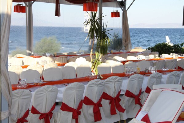 Mesa lista para una comida con sillas blancas y cintas rojas, con vista al mar.