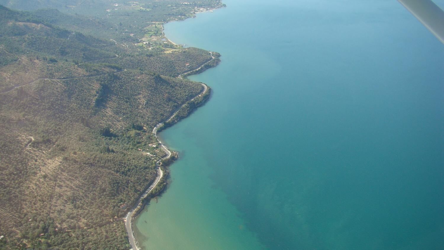 An aerial view of a coastal area with a narrow strip of land along the water, surrounded by dense vegetation and hills.