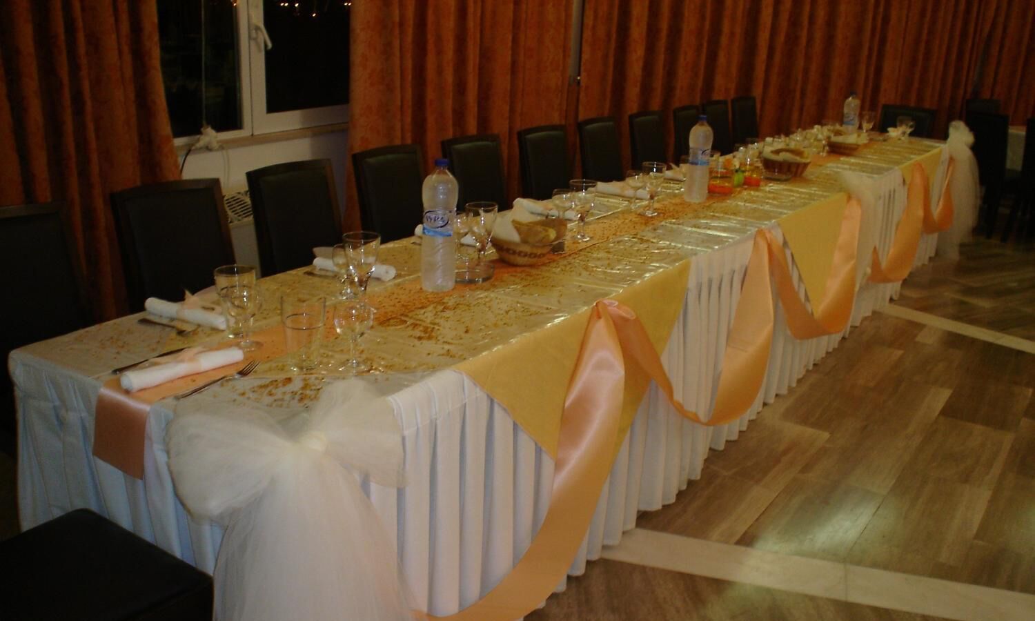 Ein langer Festtisch mit weißer Tischdecke und orangefarbenen Bändern, gedeckt mit Gläsern und Tellern, in einem Raum mit Holzboden und dunklen Vorhängen.