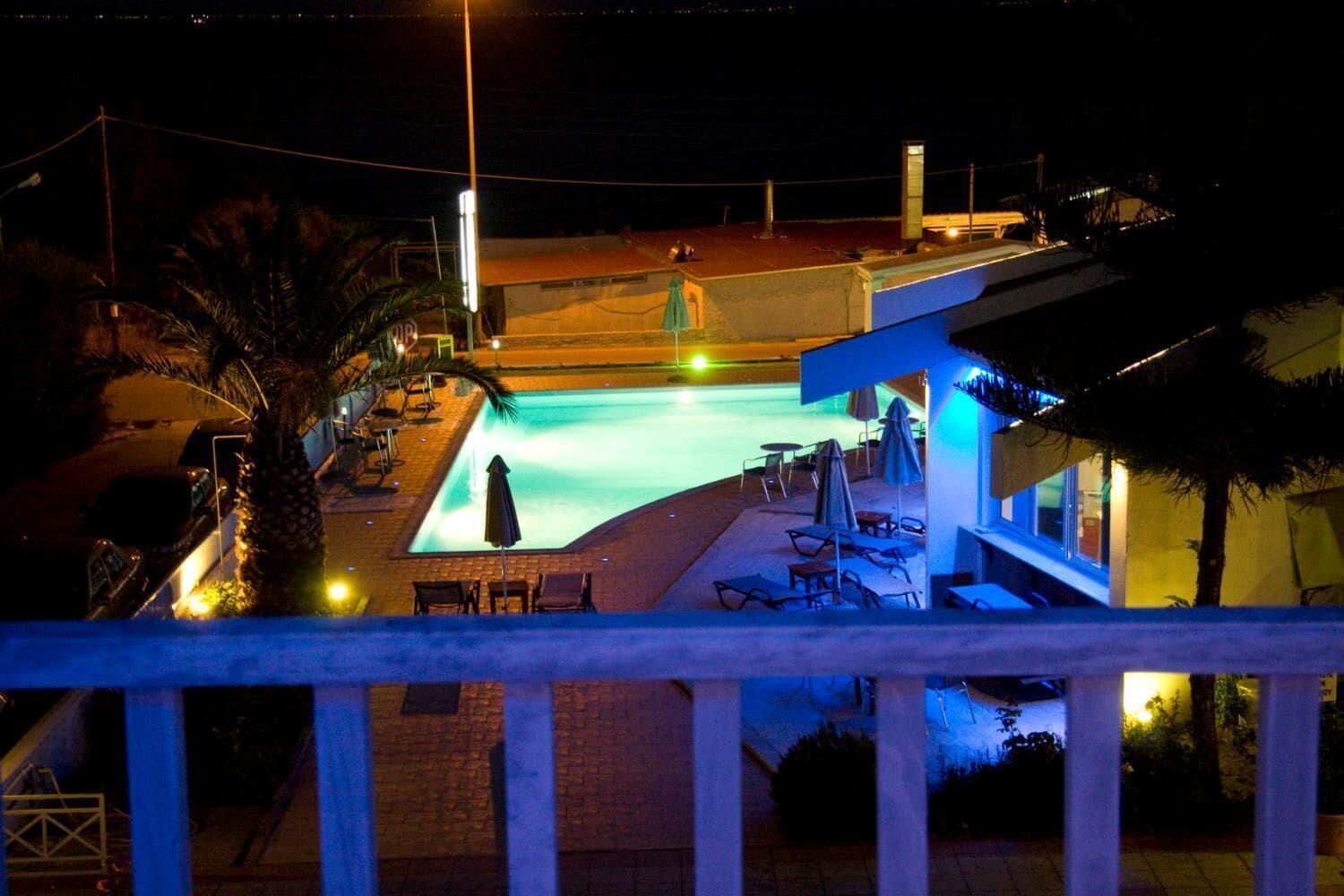 Vista nocturna de una zona de piscina con sillones de descanso y sombrillas, rodeada de palmeras y un edificio con balcón.