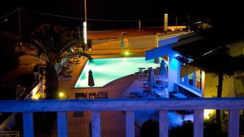 Vista nocturna de una zona de piscina con sillones de descanso y sombrillas, rodeada de palmeras y un edificio con balcón.