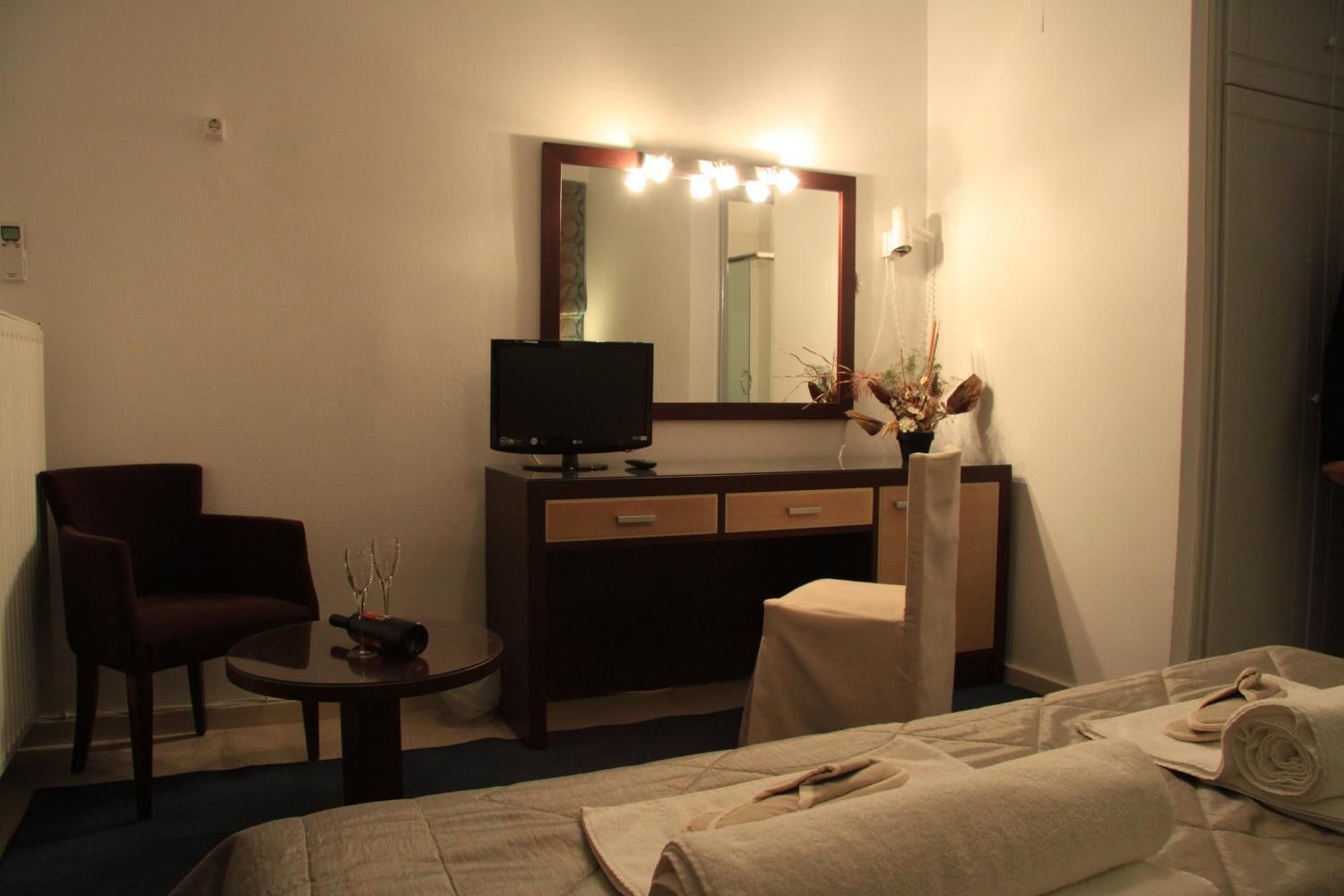 A hotel room with a bed, a desk with a computer, a chair, a small table, and a mirror with lights.