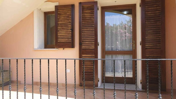 A balcony with a wrought iron railing, a door with wooden shutters, and a window with a view of trees and a clear sky. - Aria di Vacanza - Zenzero (Olbia)