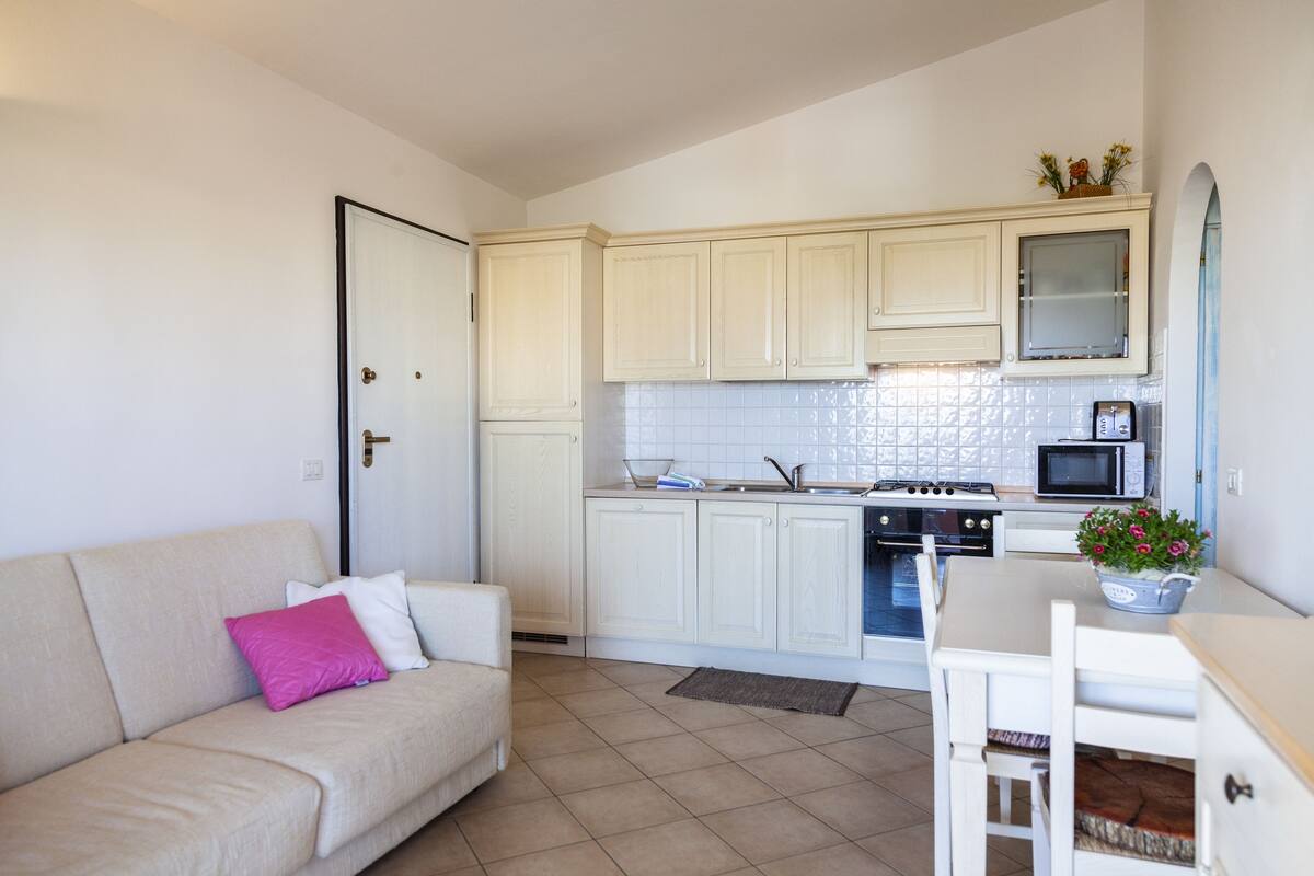 A compact kitchen with white cabinets, a microwave, and a stove. There's a beige sofa with a pink pillow and a dining table with a potted plant.