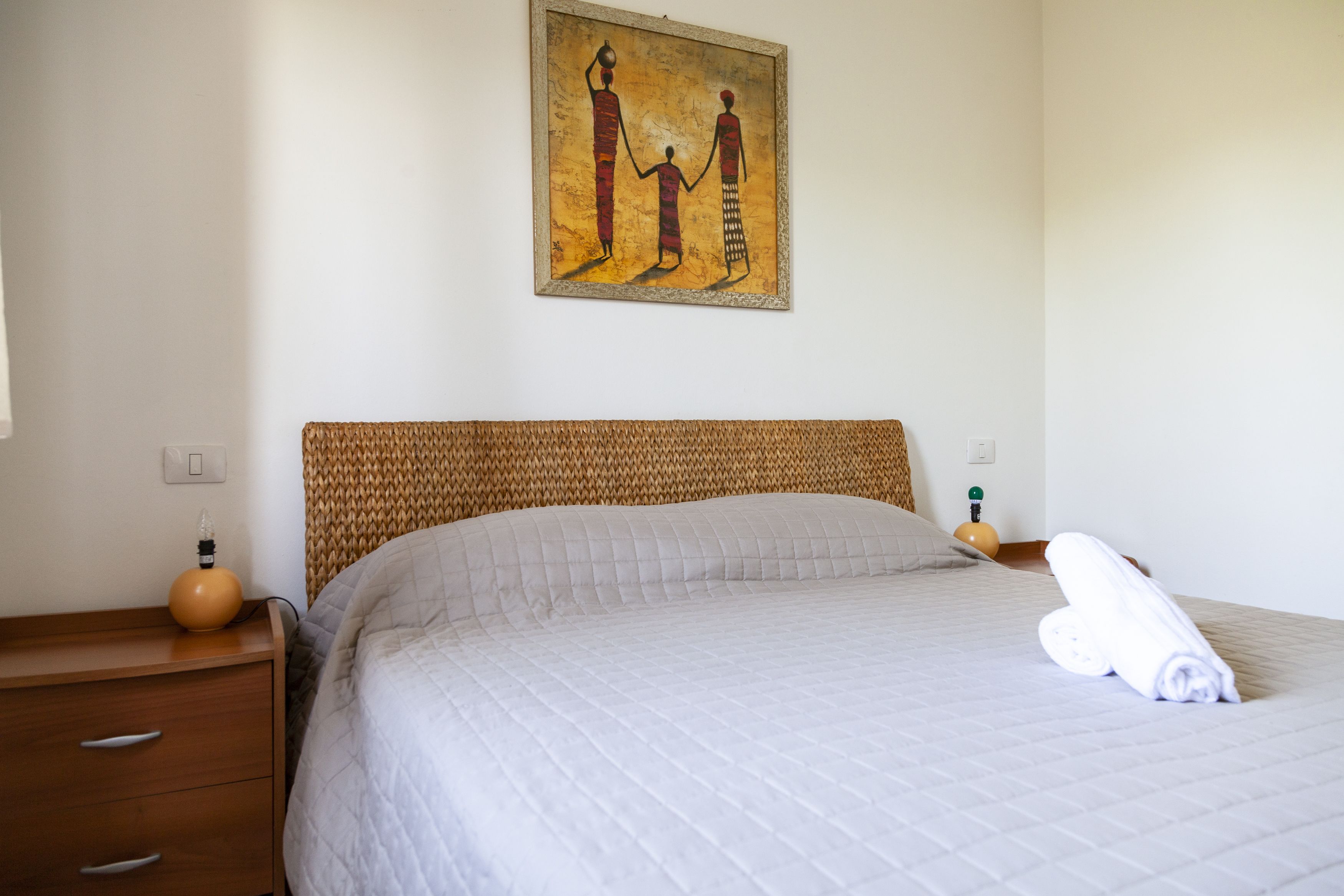 Un lit bien fait, avec une couverture matelassée, une tête de lit et un tableau représentant trois personnages accroché au mur.