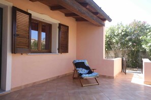 A covered patio with a single wooden chair, a window with shutters, and a view of a garden. - Aria di Vacanza - Zenzero (Olbia)