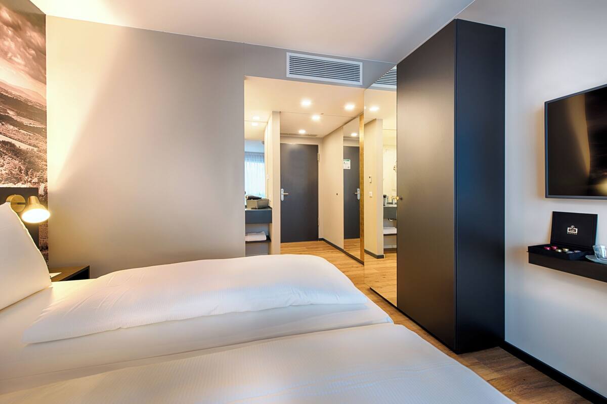 A modern hotel room with a large bed, a flat-screen TV, a bedside table with a lamp, and a view of the hallway.