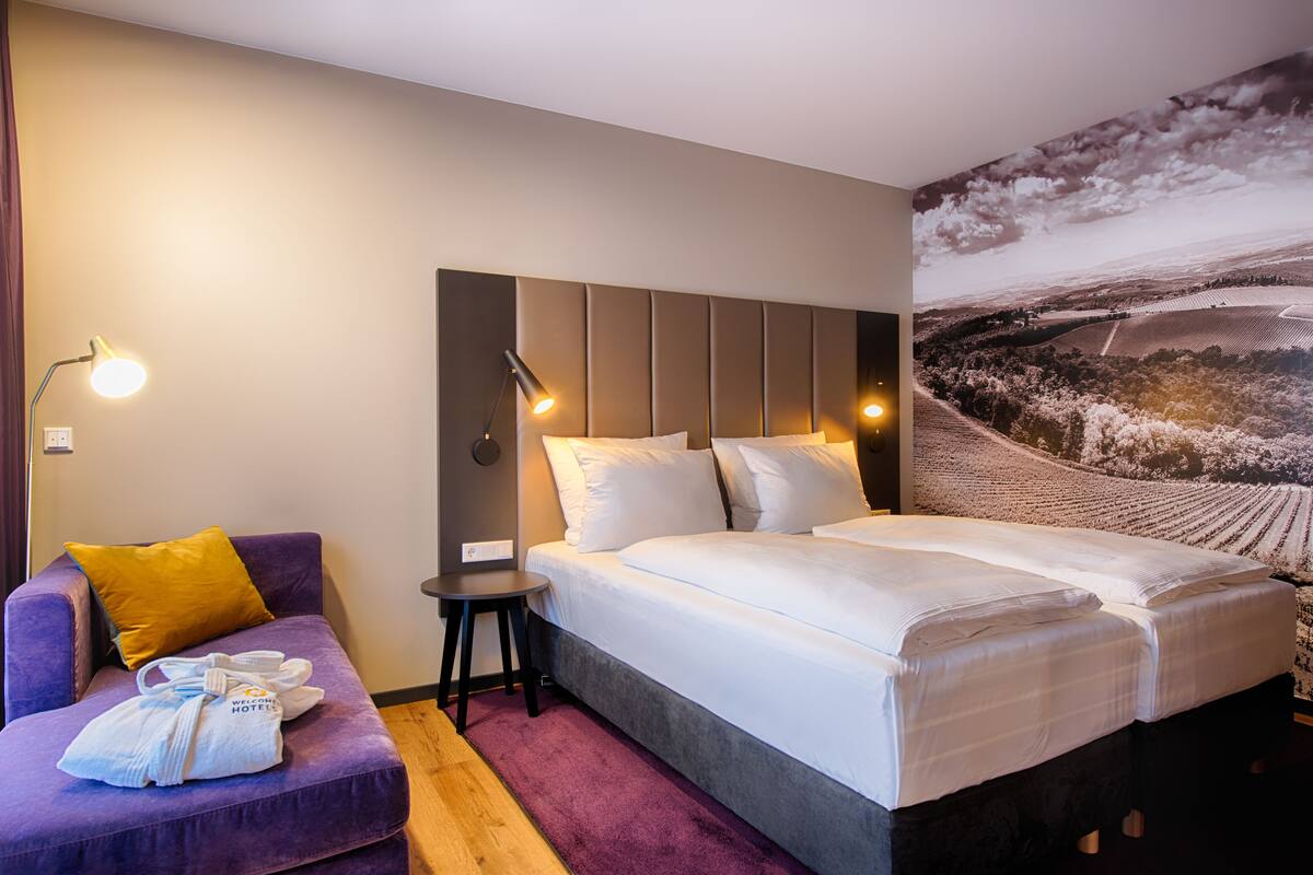 A hotel room with a bed, a purple sofa, a small table, and a wall-mounted lamp.