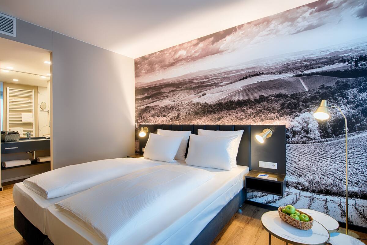 A modern hotel room with a large bed, a nightstand with a lamp, a small table with a fruit bowl, and a large landscape painting on the wall.