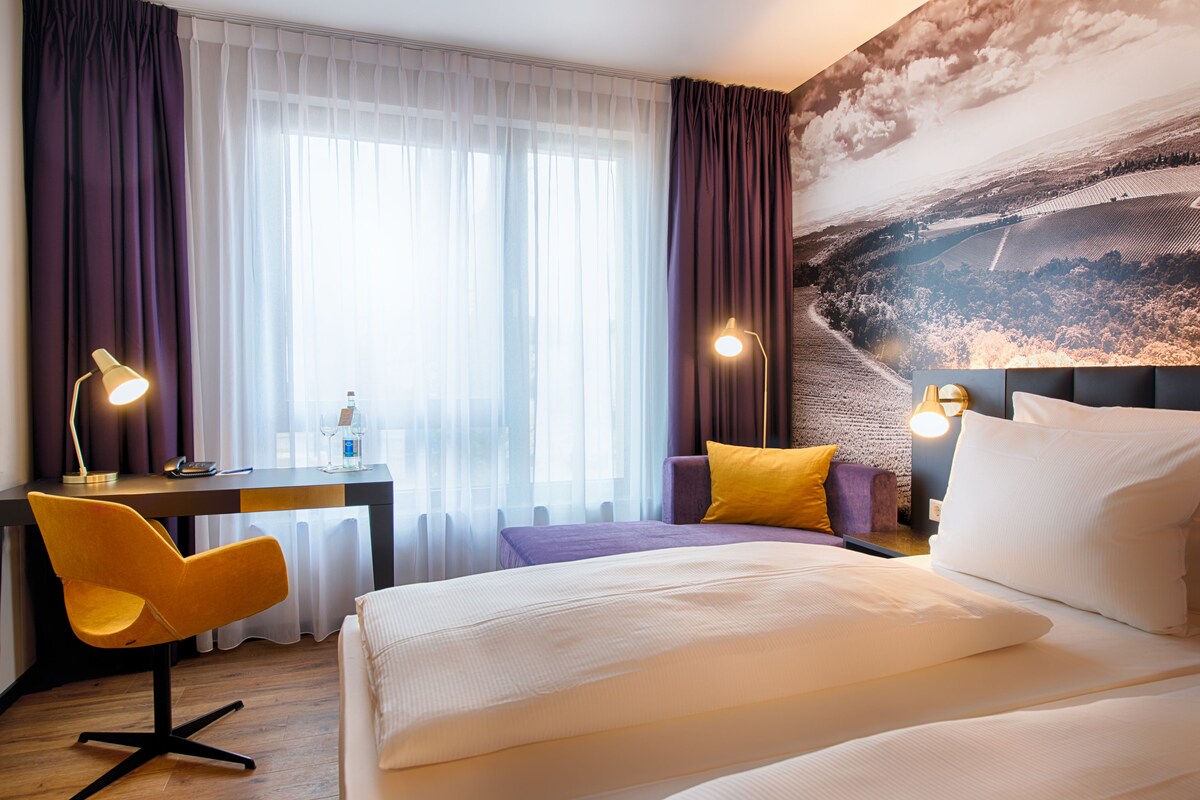 A hotel room with a bed, a desk, a chair, and a large landscape painting on the wall.