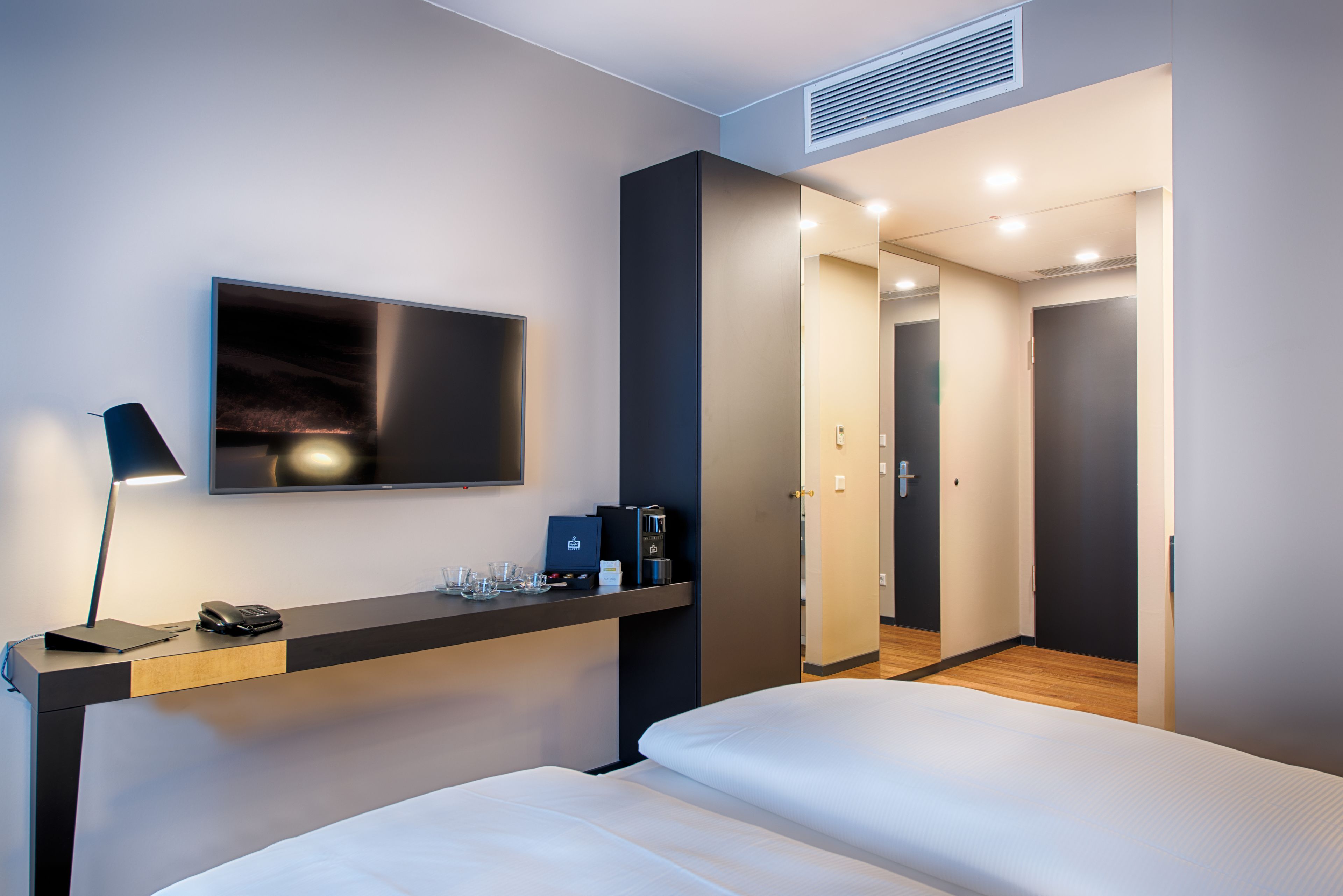 Une chambre d’hôtel moderne équipée d’un lit, d’un bureau avec une lampe, d’un téléviseur à écran plat et d’une porte donnant sur une autre pièce.