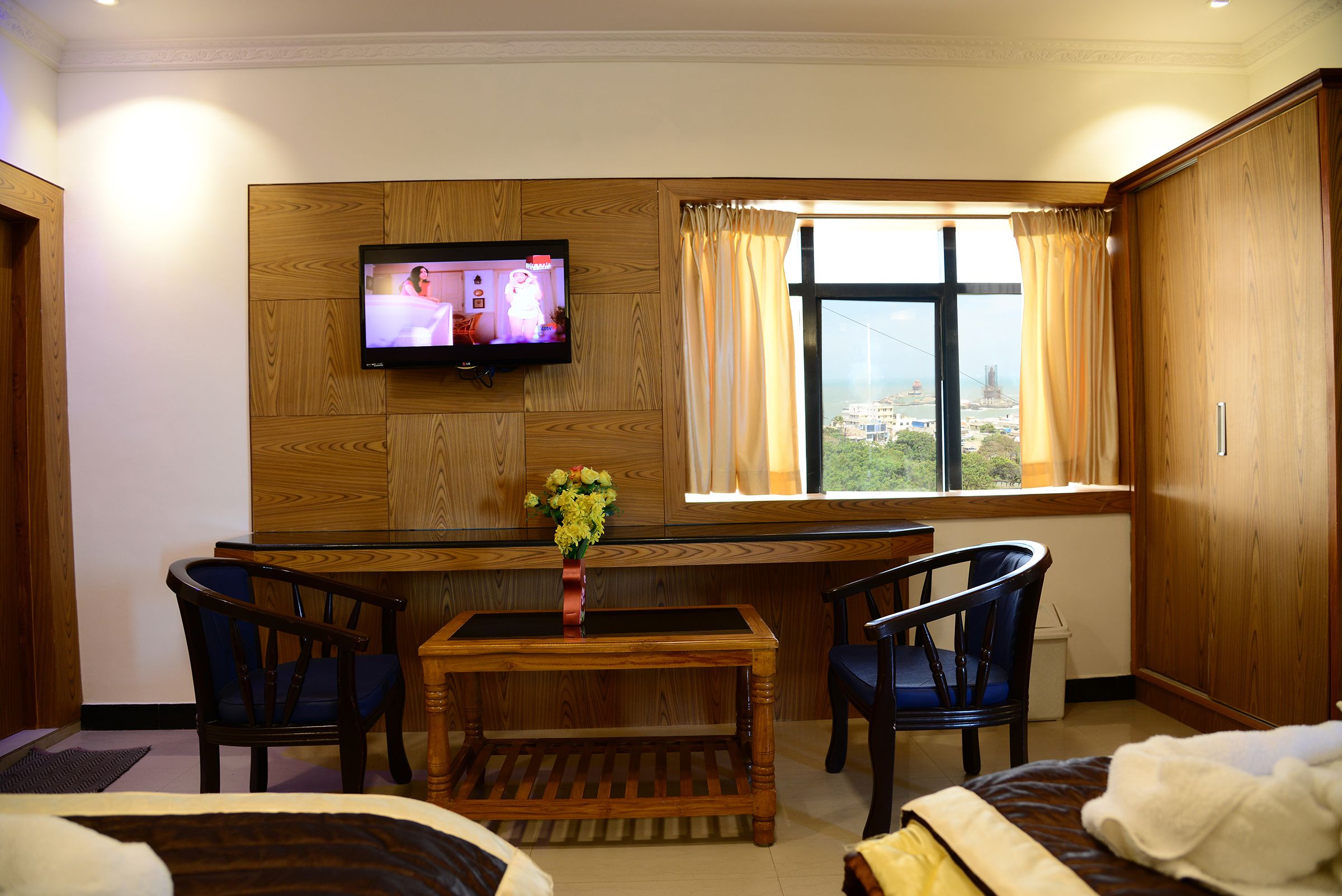 A hotel room with a wooden desk, two chairs, a TV mounted on the wall, a window with curtains, and a bed.