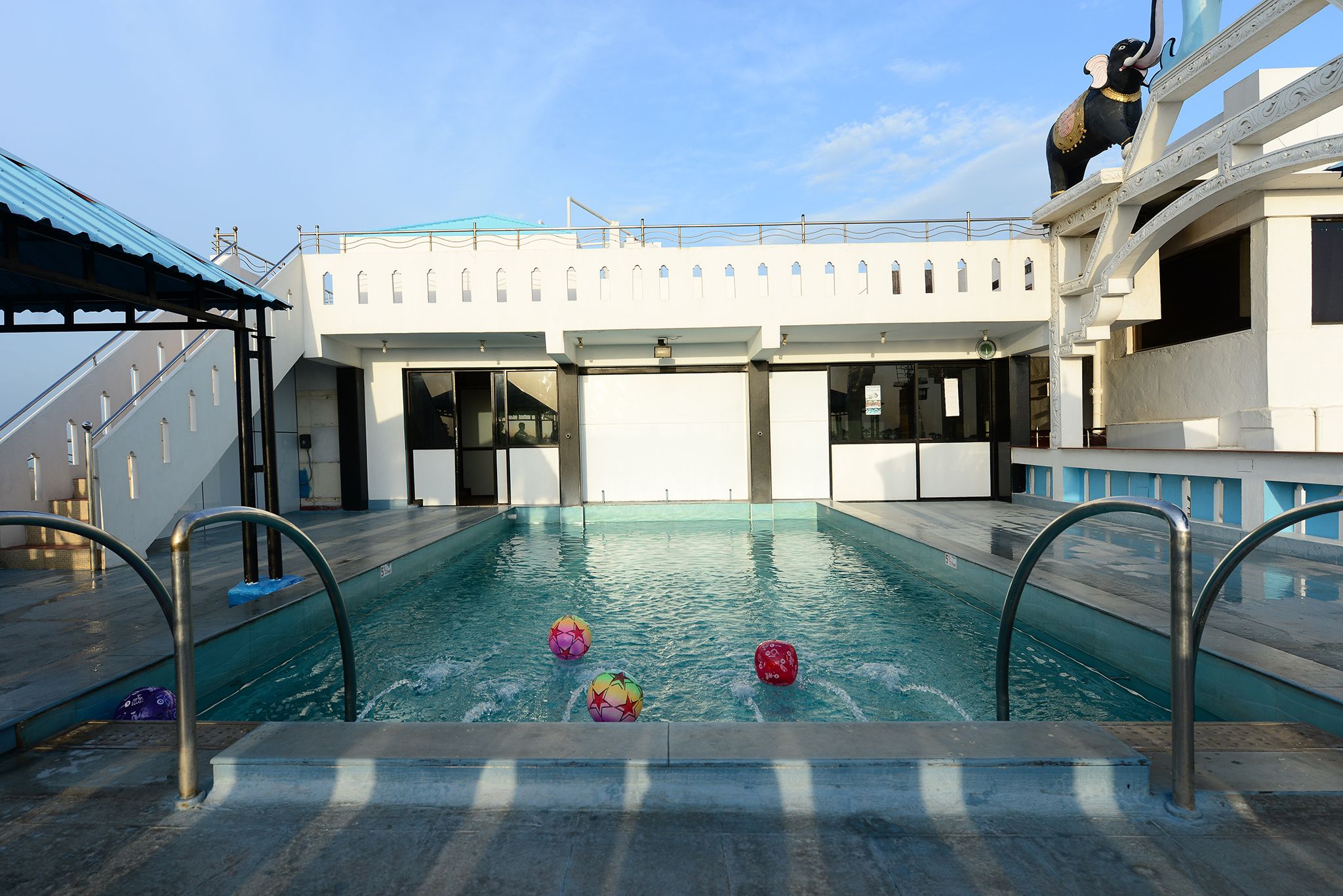 Zona de piscina con tres bolas de colores, una baranda metálica y una estatua de elefante sobre un pedestal.