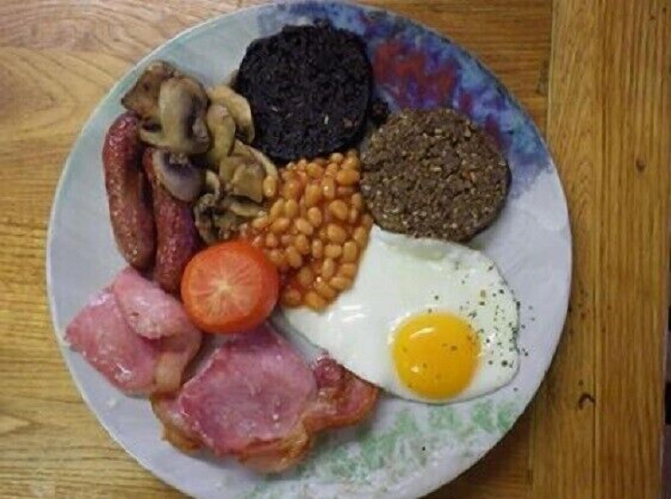 Ein Teller mit verschiedenen Frühstücksangeboten, darunter Eier, Speck, Würstchen, Pilze, Bohnen und Blutwurst.