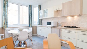 A modern kitchen with white cabinets, a microwave, and a gas stove. There is a dining table with chairs and a window with curtains. - Clos 1 (De Panne)