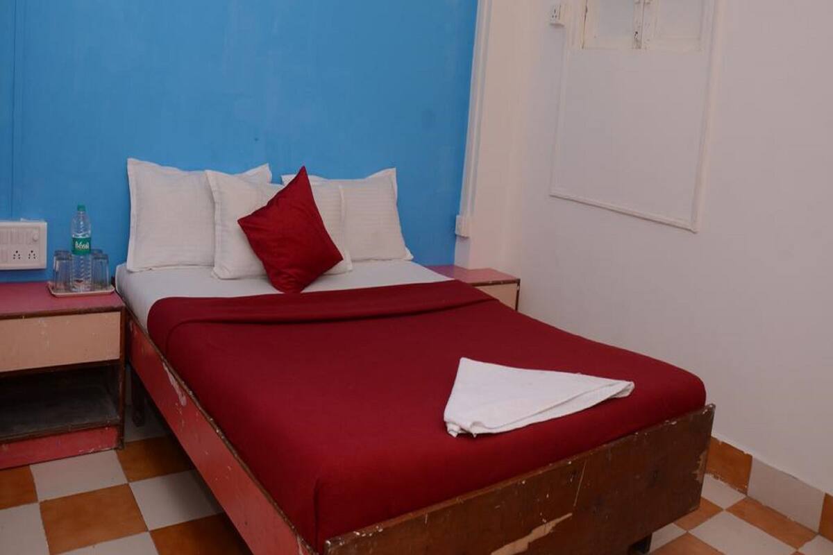 A single bed with a red bedspread and white pillows, a small bedside table with a water bottle, and a window with closed shutters.