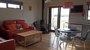 A cozy living room with a leather sofa, wooden coffee table, and a dining area with a table and chairs. A fireplace is visible in the background, and there is a large window providing a view of the outdoors. - Tor Farm Lodge (Cheddar)