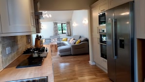 A modern kitchen with a built-in stove, a refrigerator, and a dining area with a wooden table and a grey sofa. - Tor Farm Lodge (Cheddar)