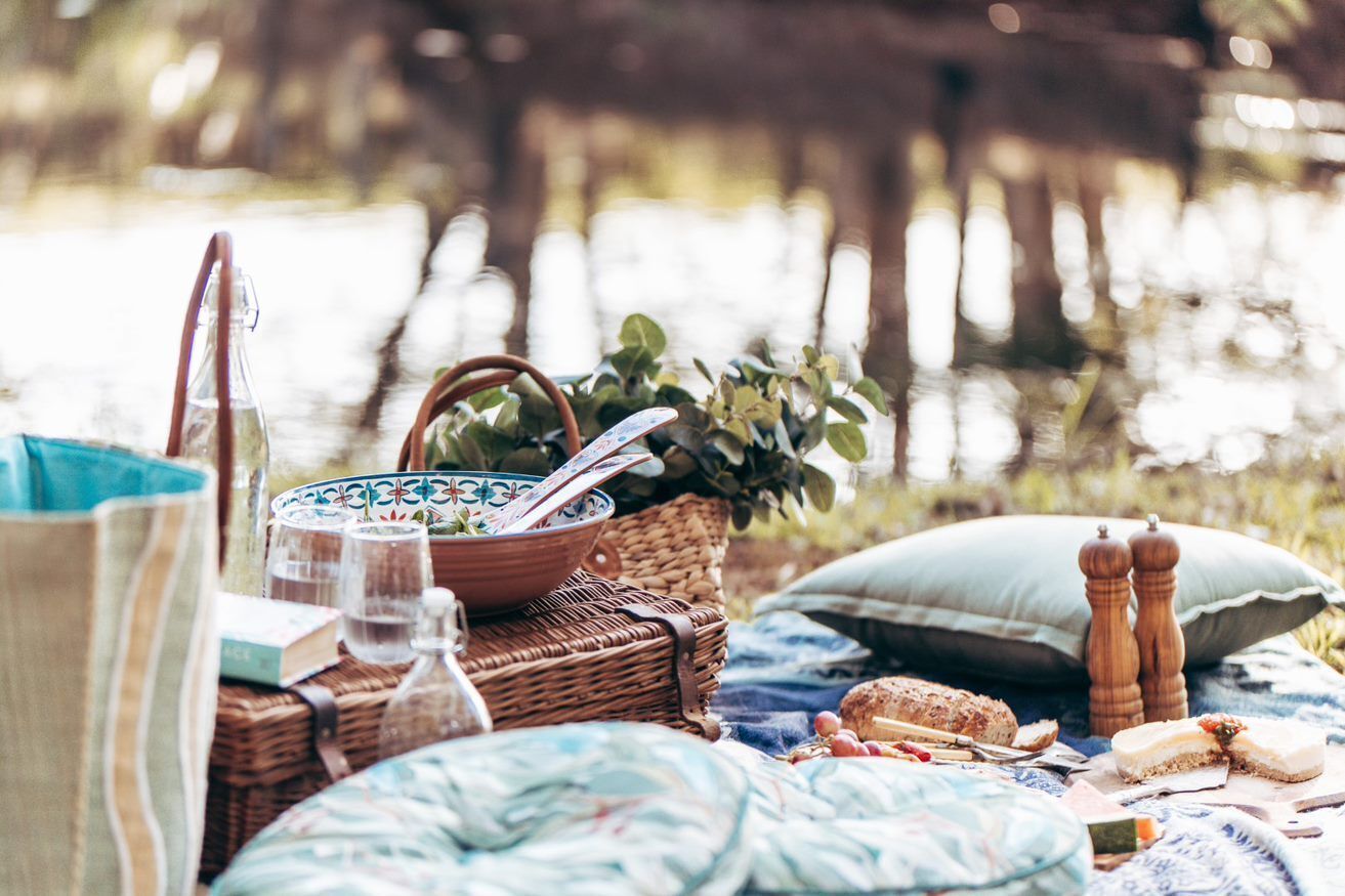 Een picknickopstelling met een mand, een schaal, een boek en een fles op een deken bij het water.