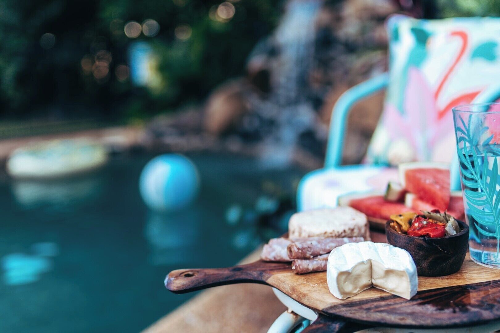 A dining table with a wooden cutting board, a cheese wheel, a bowl of fruit, and a glass of beverage.