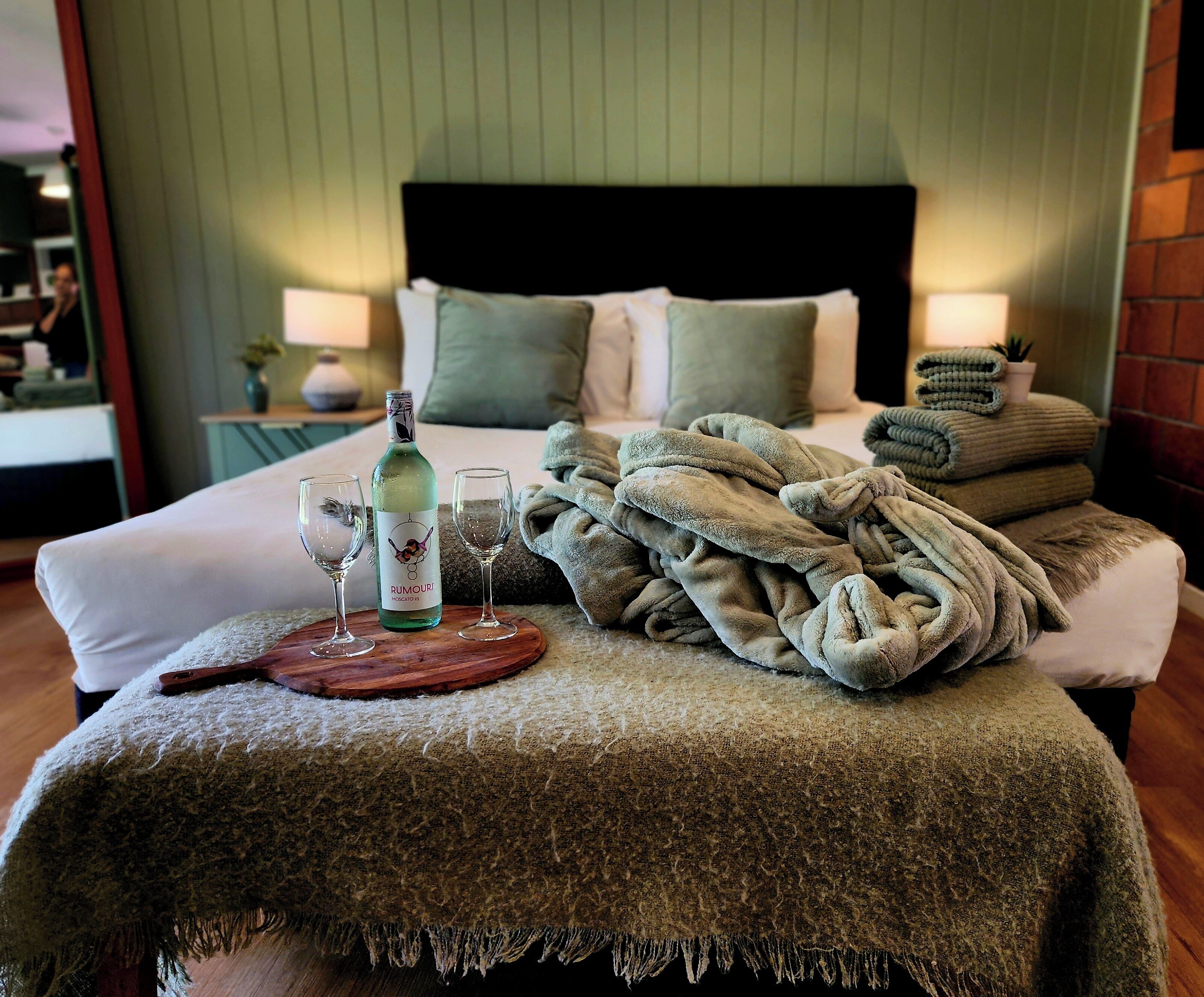 A neatly made bed with a green blanket, a bottle of wine, and two glasses on a wooden tray.