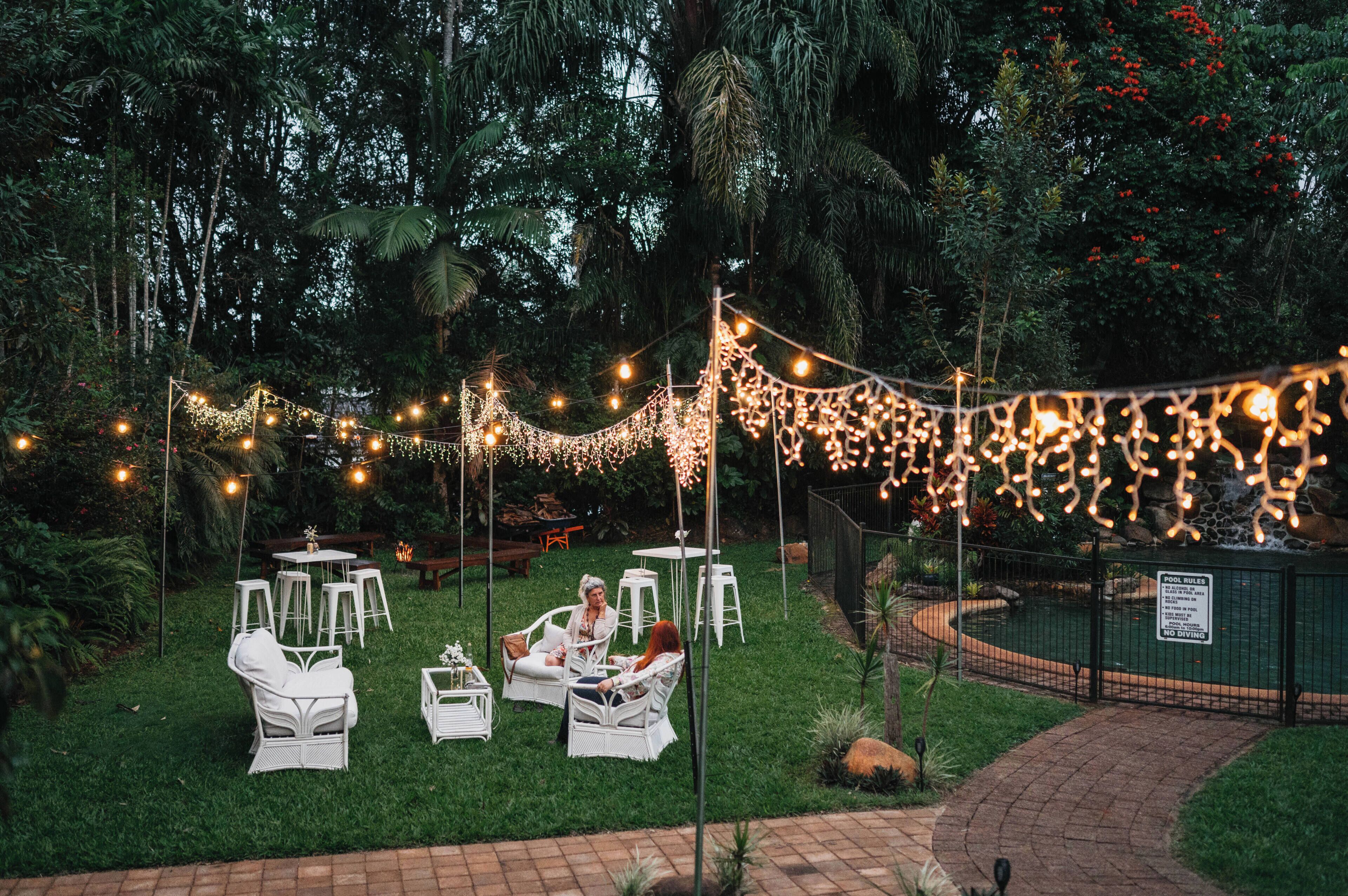 An outdoor seating area with white chairs and tables, surrounded by greenery and string lights.