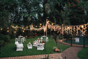 Un espacio al aire libre con sillas y mesas blancas, rodeado de follaje y luces colgantes.