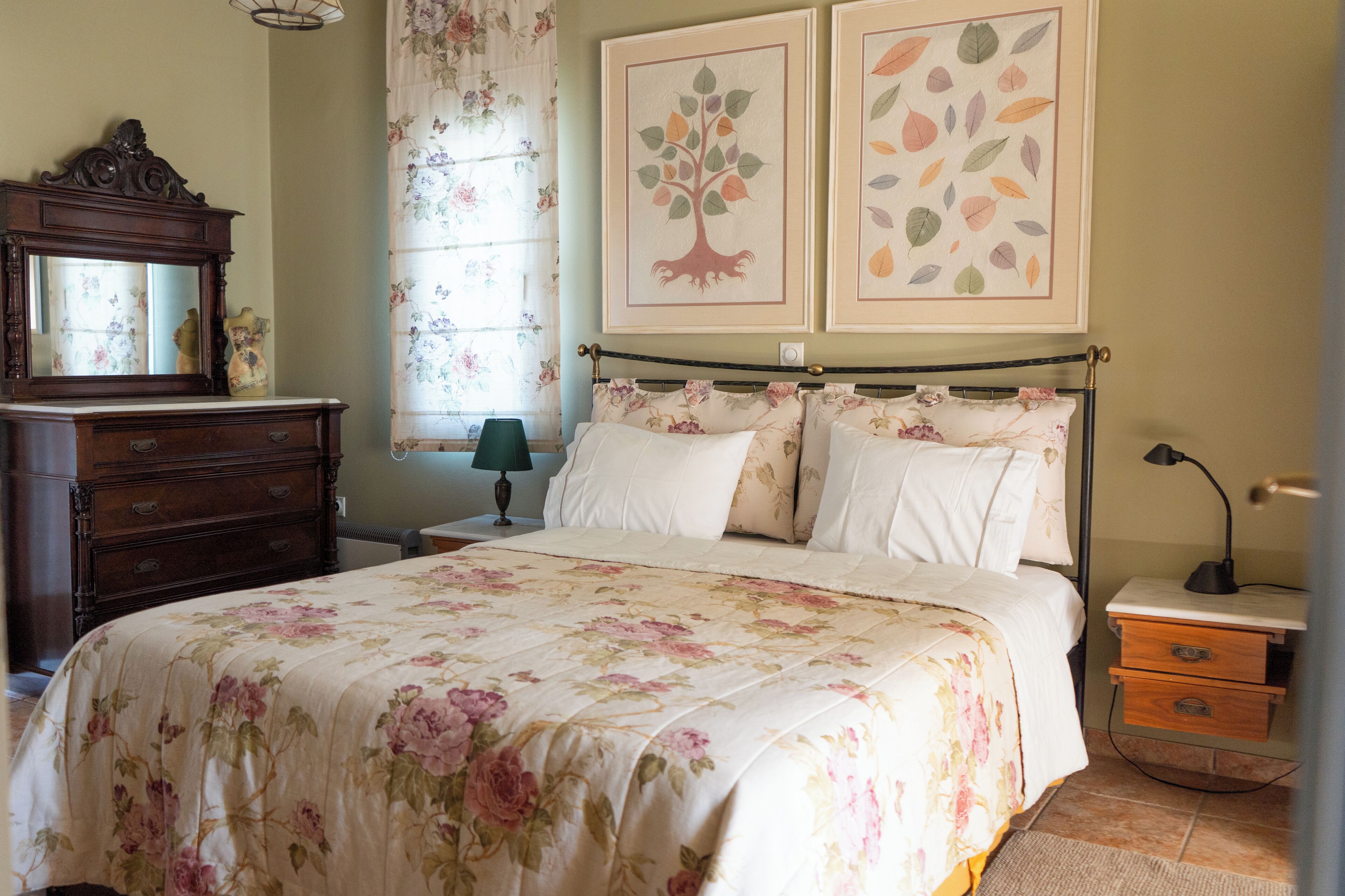 A bedroom with a floral bedspread, a wooden dresser, a nightstand with a lamp, and two framed pictures on the wall.