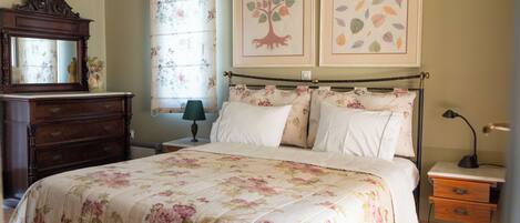 A bedroom with a floral bedspread, a wooden dresser, a nightstand with a lamp, and two framed pictures on the wall.