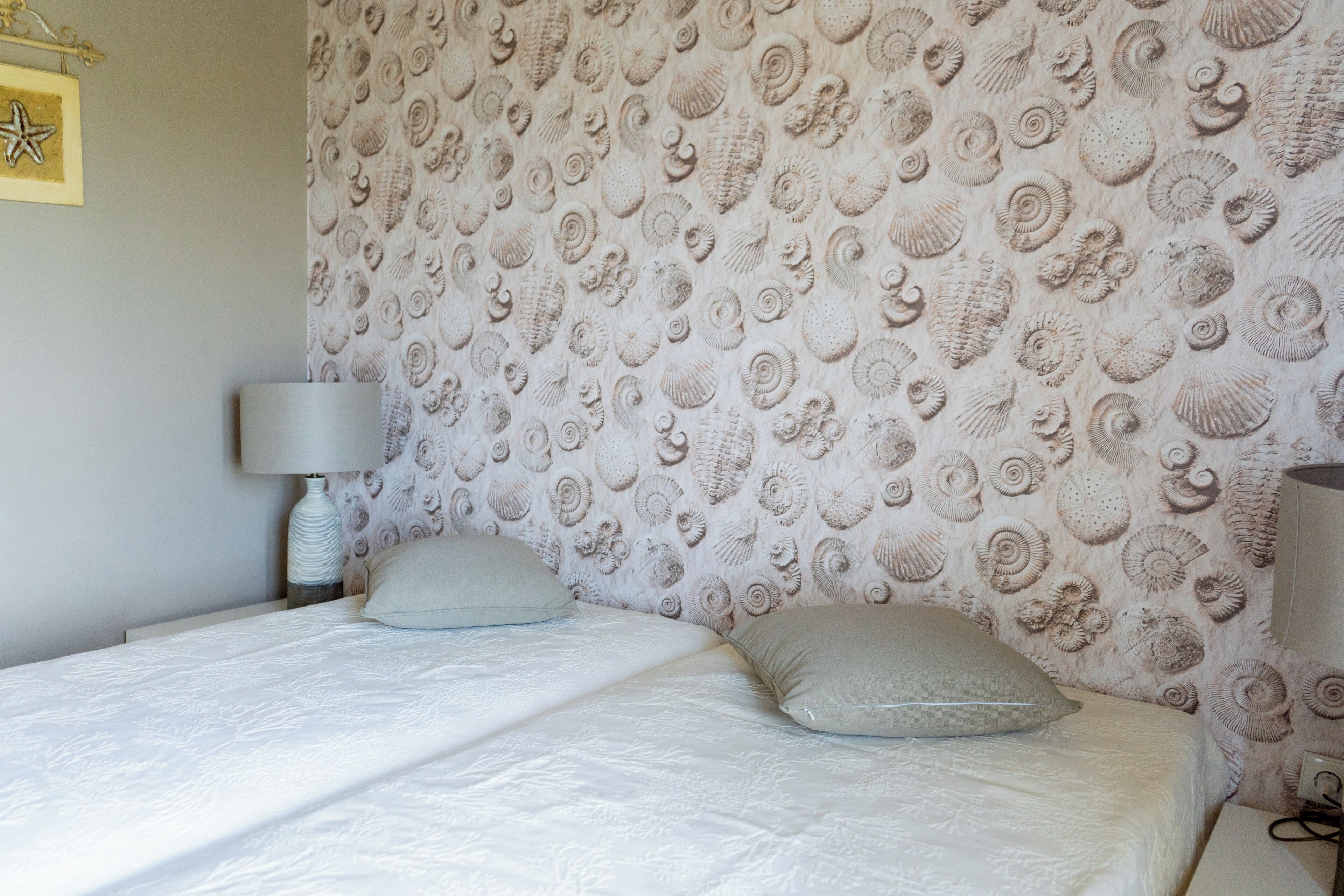 A bedroom with a bed, two pillows, a bedside lamp, and a wall with a shell pattern.