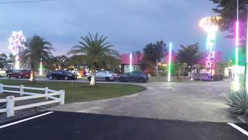Un estacionamiento con coches y palmeras, cerca hay una valla blanca y luces de colores.