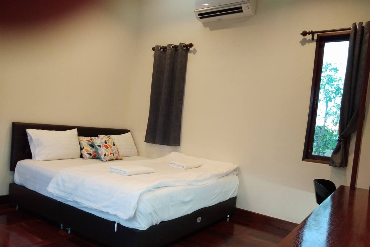 A neatly made bed with white linens and a colorful pillow, a wooden headboard, a window with curtains, and an air conditioning unit on the wall.